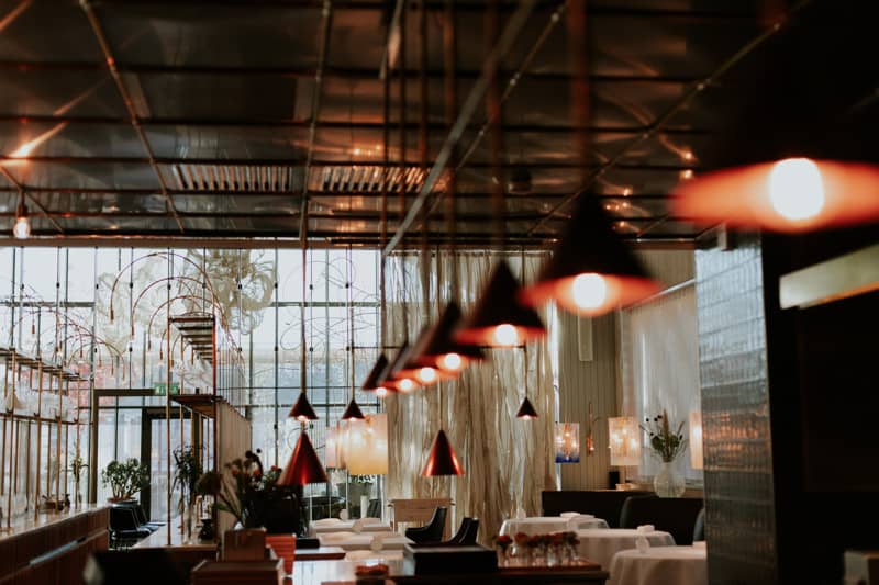 Warm pendant lights hang in rows over an elegant restaurant interior, with white tablecloths, flowers, and large windows letting in daylight behind sheer curtains.