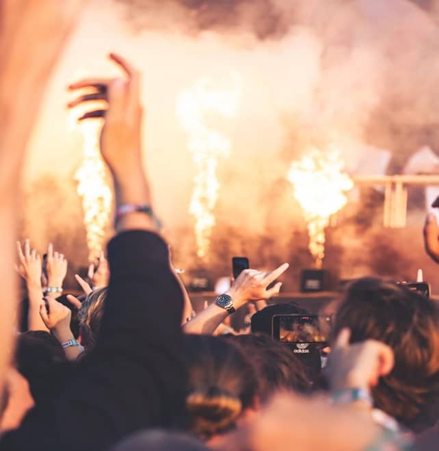 A dense crowd at a concert with raised hands and phones filming, while bright flames and smoke shoot up from the stage in the background.