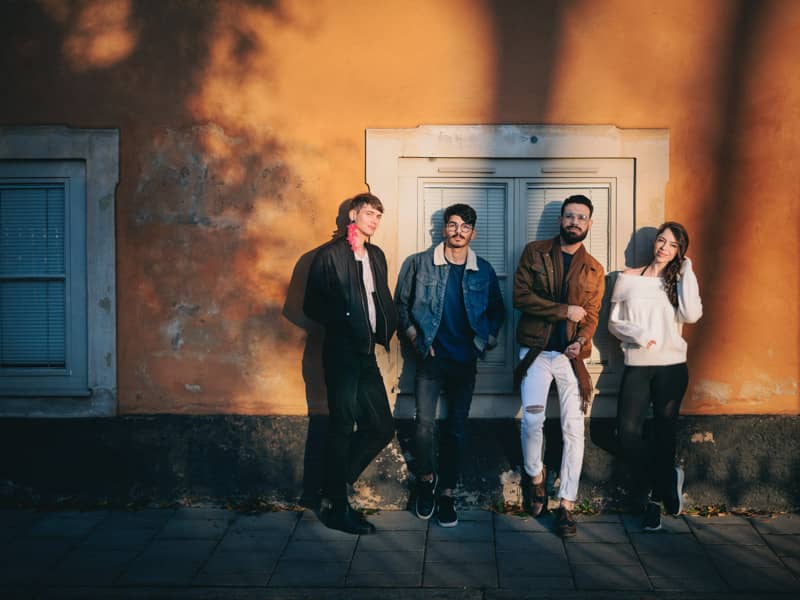Four young adults stand relaxed against a warm orange wall in sunlight, leaning and smiling toward the camera on a city sidewalk.
