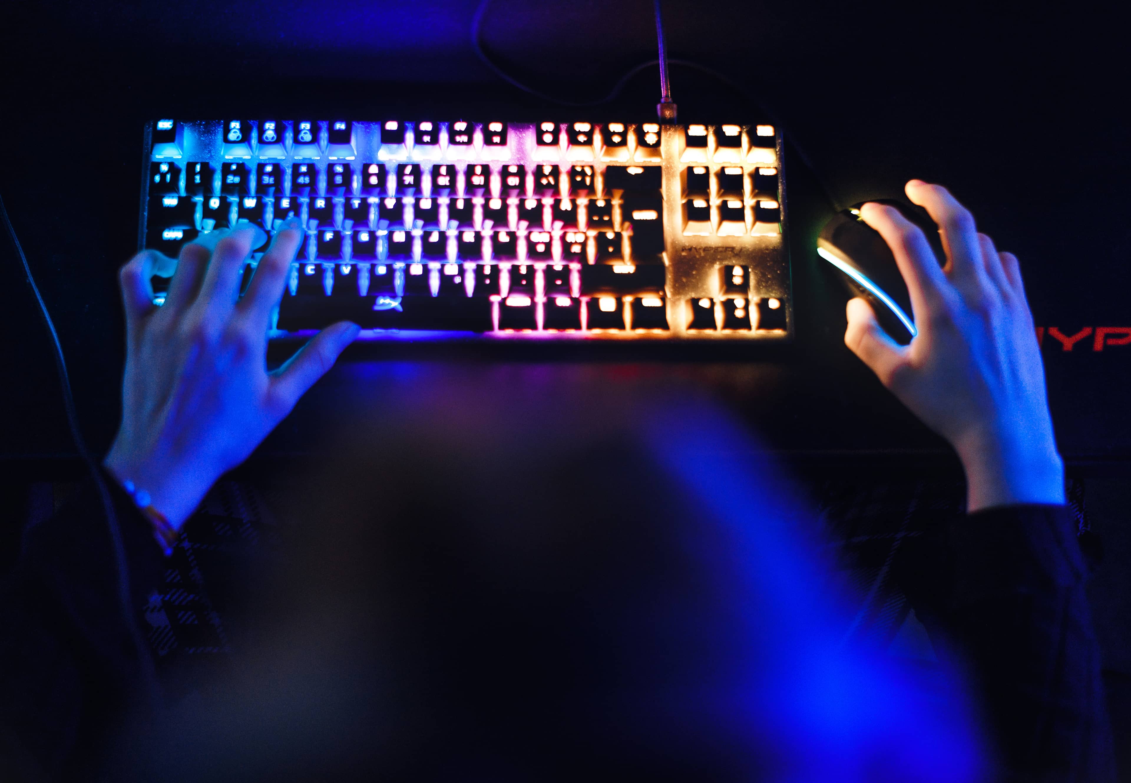 Hands on a brightly lit rainbow keyboard and mouse in a dark room with blue light.
