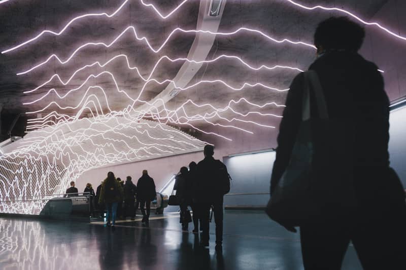 People walk through an underground corridor with an escalator, under a ceiling covered in wavy neon light lines reflecting on the shiny floor.