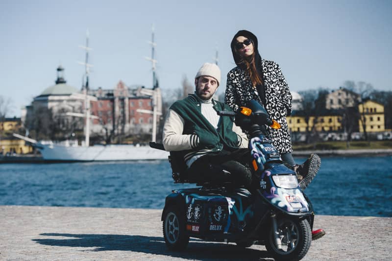A person sits on a decorated mobility scooter beside a friend standing next to them, both wearing winter clothes, with water, a white sailing ship and colorful buildings in the background.