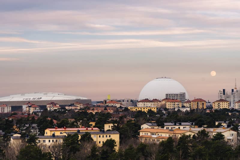 View over a residential area in southern Stockholm with a large white spherical arena and a flat-roofed stadium on the horizon, under a pastel evening sky with the moon rising.