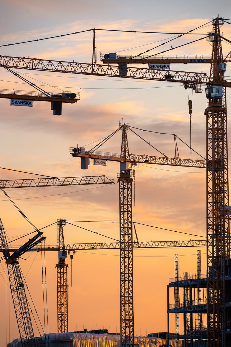 Several tall tower cranes rise above a construction site, silhouetted against an orange and pink sunset sky.