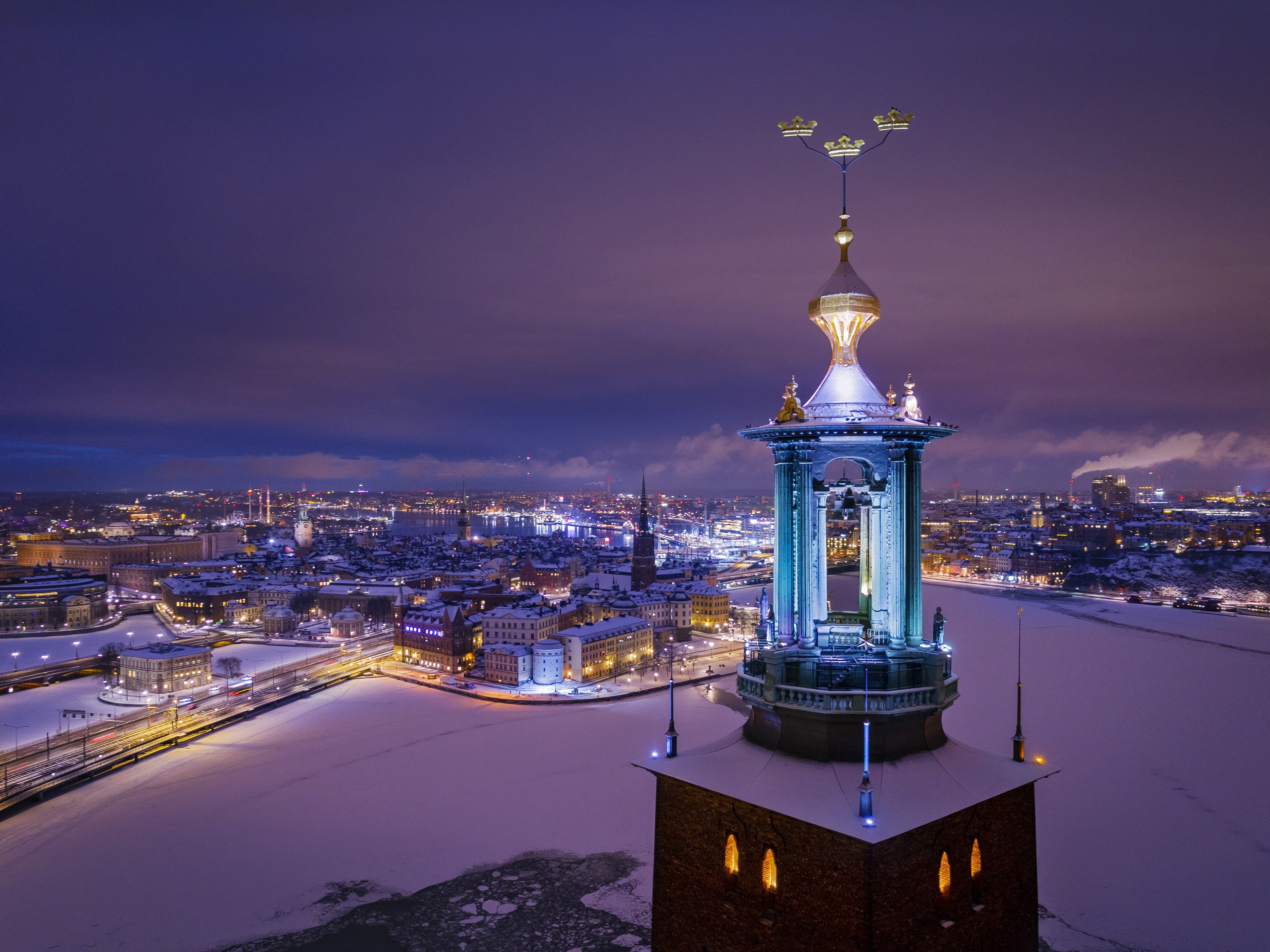 View of snow-covered Stockholm at dusk with the illuminated tower of City Hall and its three golden crowns in the foreground.