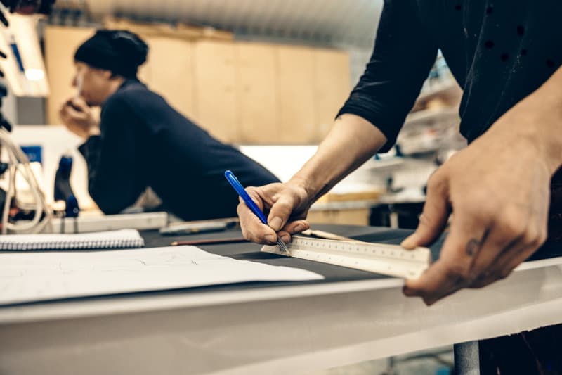 Close-up of a person drawing a straight line with a pen and ruler on a worktable, with sketches and tools nearby and another person blurred in the background of the workshop.