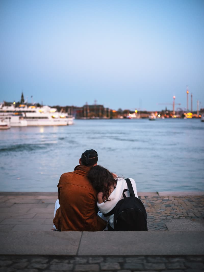 Two people sitting close together on the quay, one resting their head on the other’s shoulder, with water and boats in the dusk light.