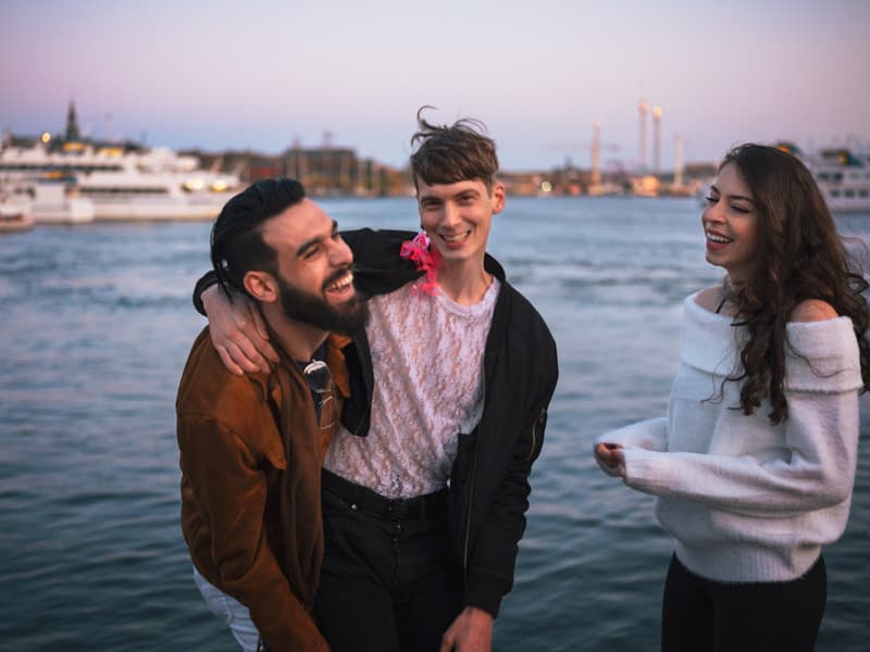 Three people stand close together by the water, laughing and sharing a relaxed, warm moment. Two of them have their arms around each other while the third stands nearby smiling. The image conveys friendship and joy in an urban waterfront setting at dusk.