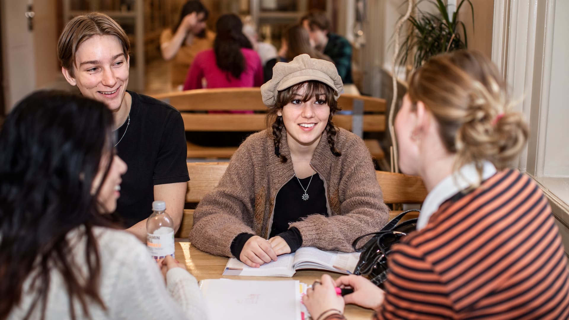 Four young people sit around a table talking, with books and notes in front of them. They smile and engage with each other in a warm, social setting. The image conveys togetherness, studying, and collaboration.