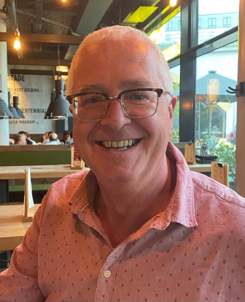 A smiling person with glasses and a pink patterned shirt sits at a wooden table inside a lively restaurant, with other diners and large windows in the background.