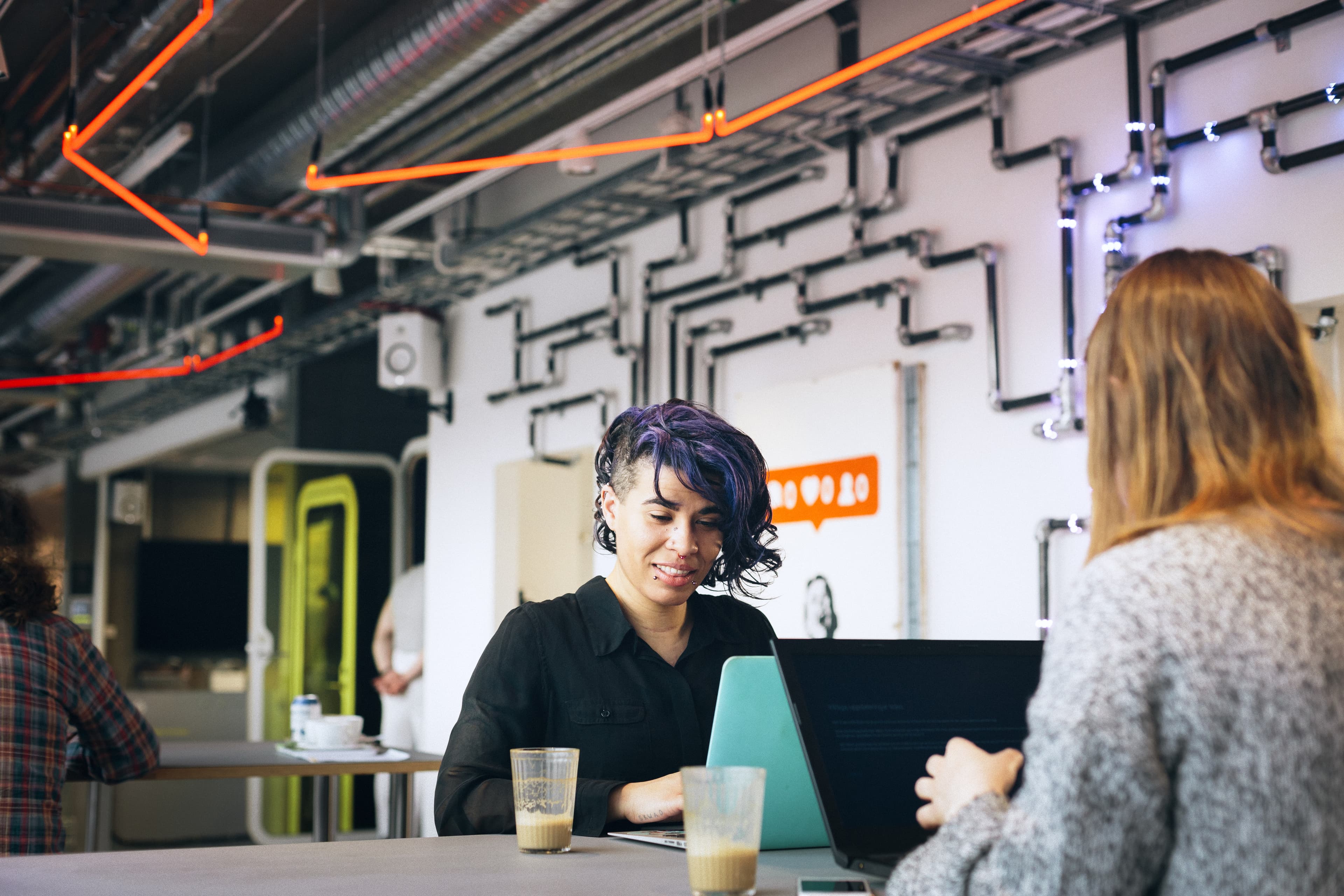 Två personer sitter vid ett bord med laptops och kaffe i en modern lokal med neonljus och industriell inredning.
