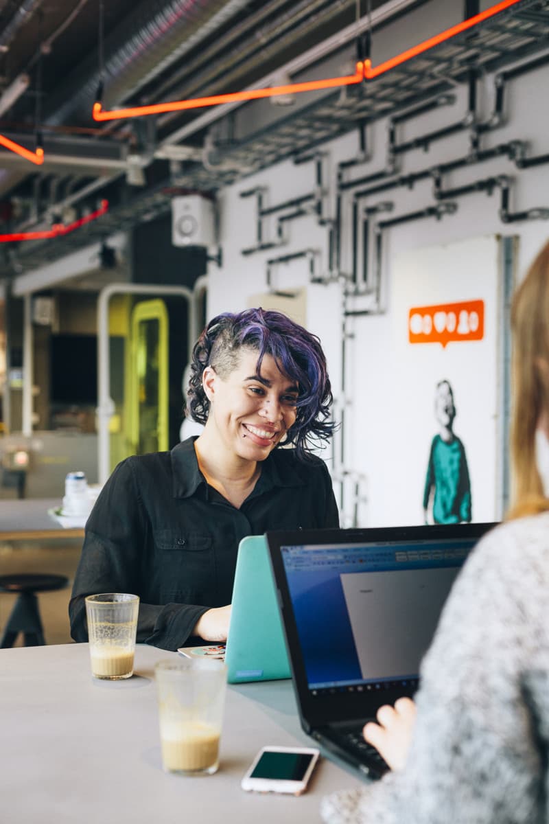 Smiling person with purple hair working on a laptop in a modern office, with a coworker blurred in the foreground.