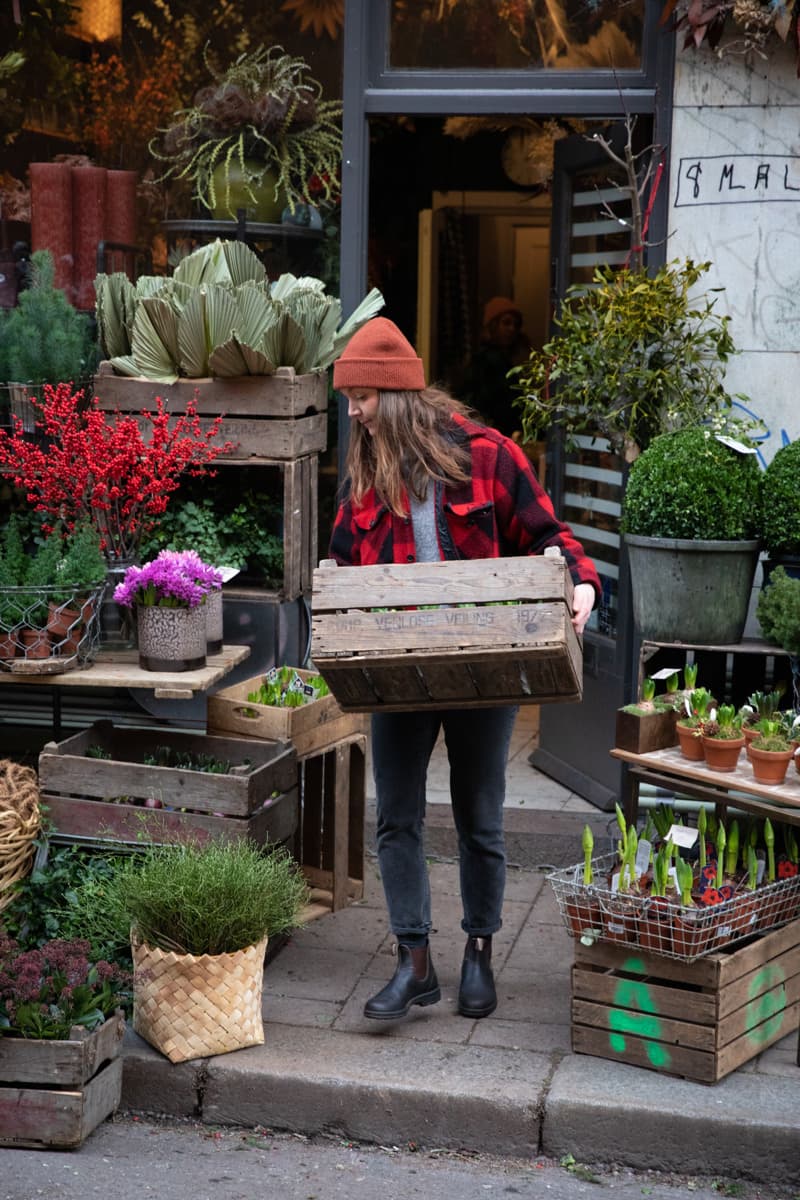 Person in a checkered jacket and beanie carrying a wooden crate in front of a flower shop with plants and flowers displayed on the sidewalk.