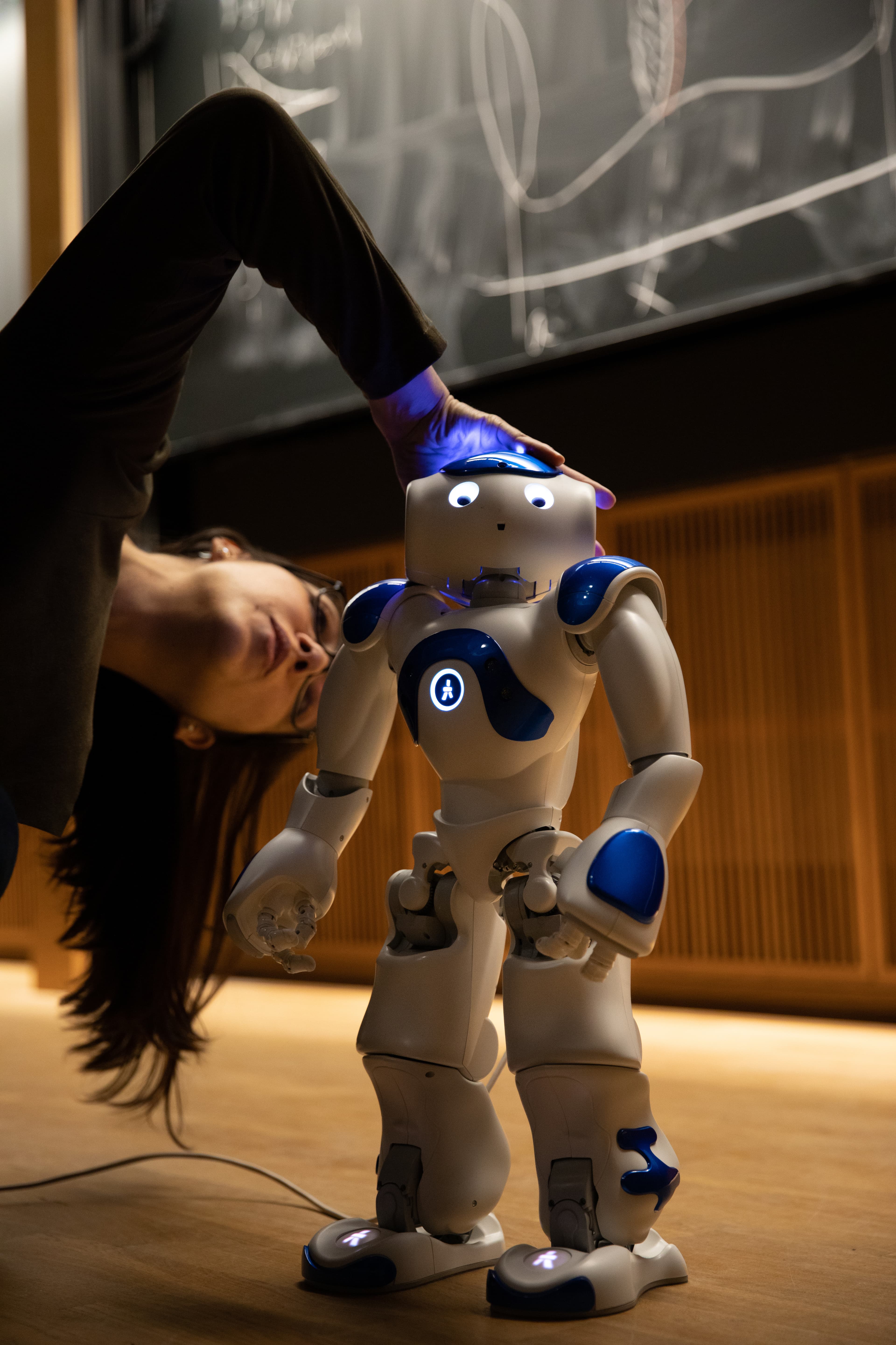Person leaning down to adjust a small white and blue humanoid robot in front of a chalkboard in a lecture hall.