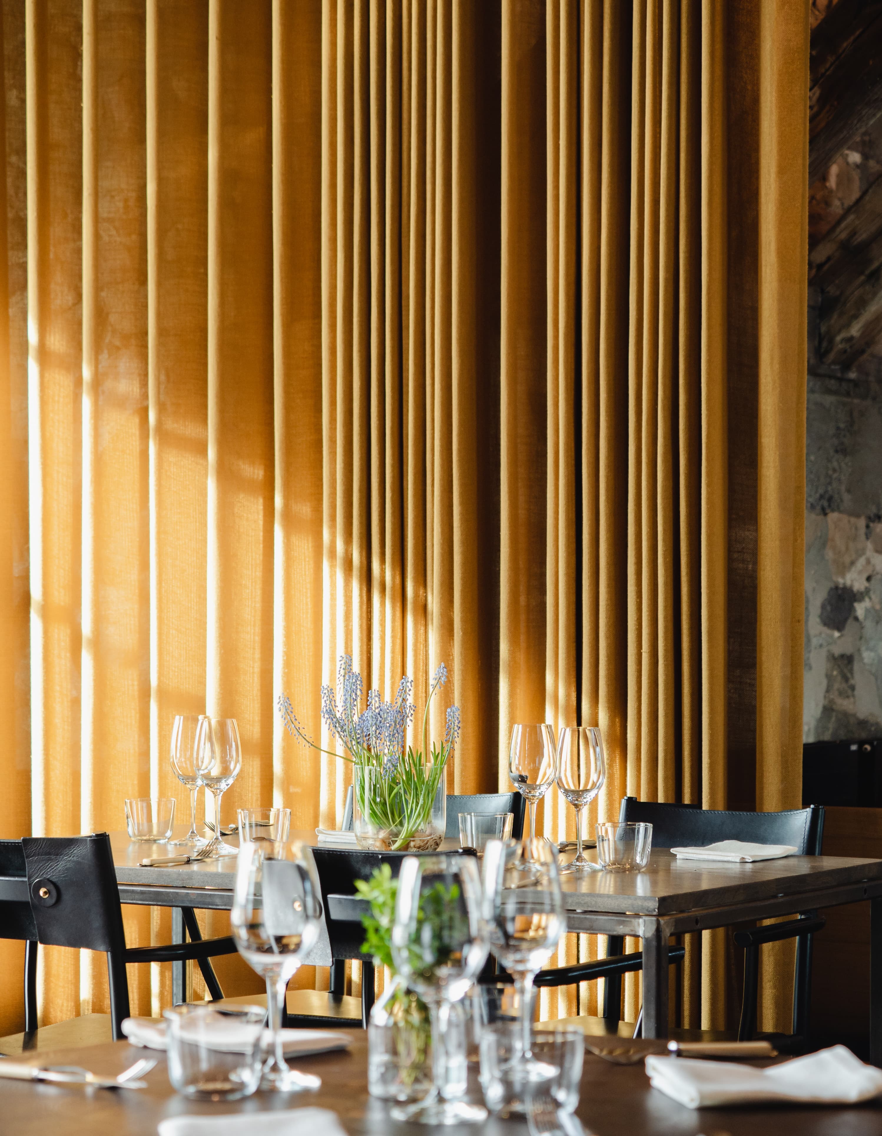 Set restaurant table with wine glasses and flowers in front of tall golden curtains in soft daylight.