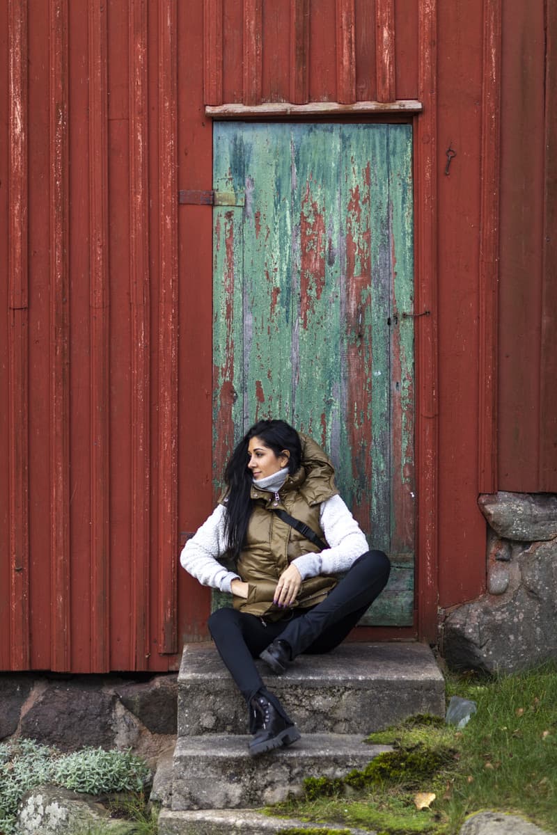 Person in a warm vest sitting on stone steps in front of a red wooden wall with a weathered green door in a coastal countryside setting.