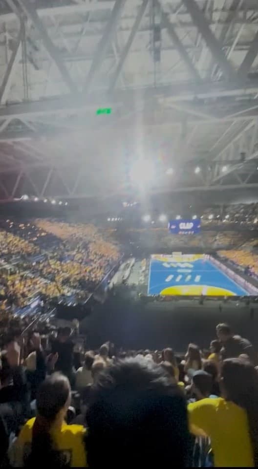Large indoor arena with a blue court and fans in yellow shirts standing and cheering.