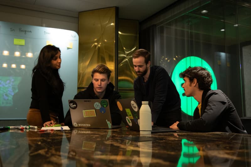 Four colleagues gather around a conference table with laptops and markers, discussing work in a modern glass-walled office with a whiteboard full of sticky notes in the background.