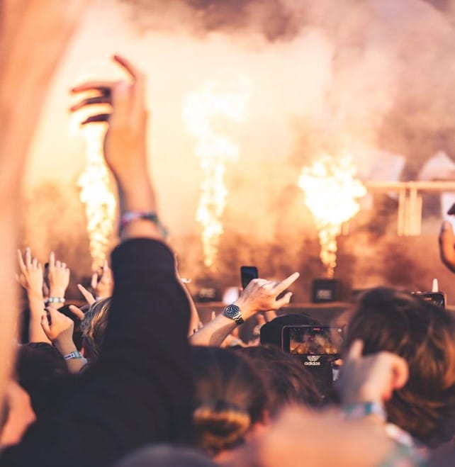 A dense crowd at a concert with raised hands and phones filming, while bright flames and smoke shoot up from the stage in the background.