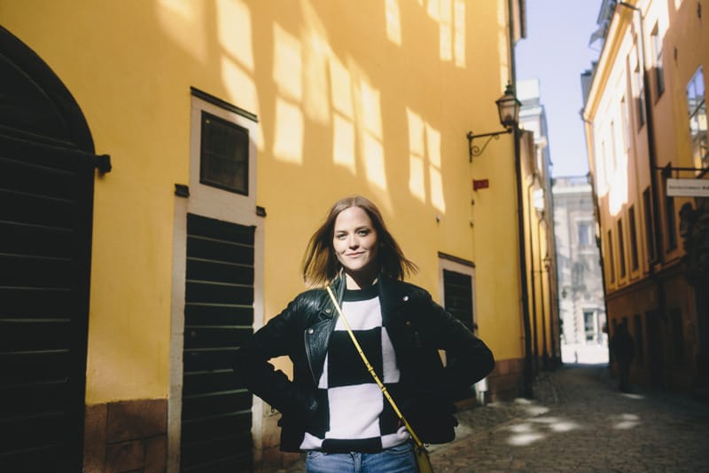 A smiling person stands with hands on hips in a narrow cobblestone street lined with bright yellow buildings, sunlight casting window-shaped shadows on the wall behind.