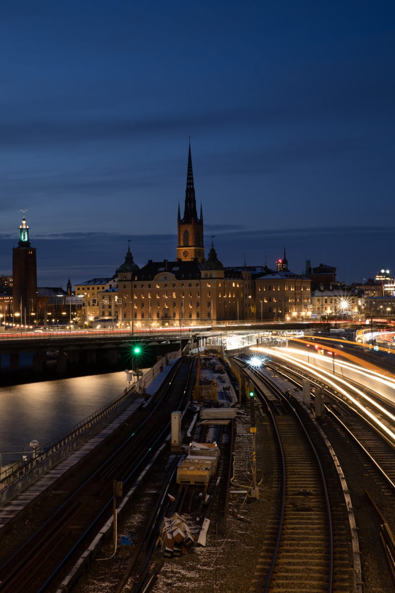Night view of central Stockholm with Riddarholmen Church lit up, Stockholm City Hall on the left, and light trails from trains and traffic curving along the railway tracks and bridge over the water.