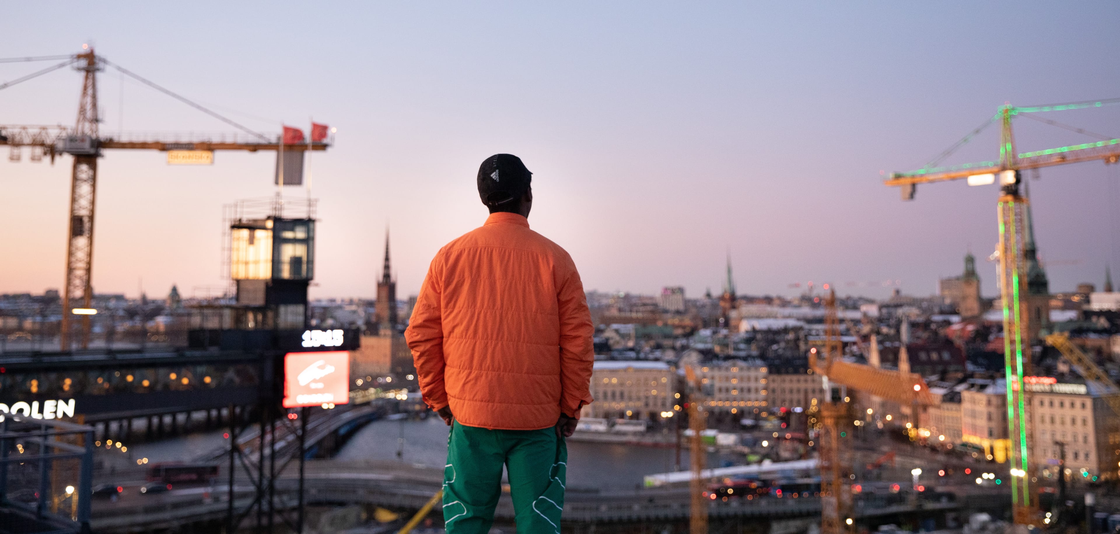 A person wearing an orange jacket and green pants stands on a high vantage point, looking out over a city at dusk, surrounded by construction cranes and city lights. The person faces away from the camera, taking in the wide urban landscape. The image conveys reflection and a sense of observing a city in transition.