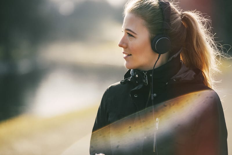 Woman in a jacket wearing headphones looking to the side while walking outdoors, with soft backlight and light flares in the image.