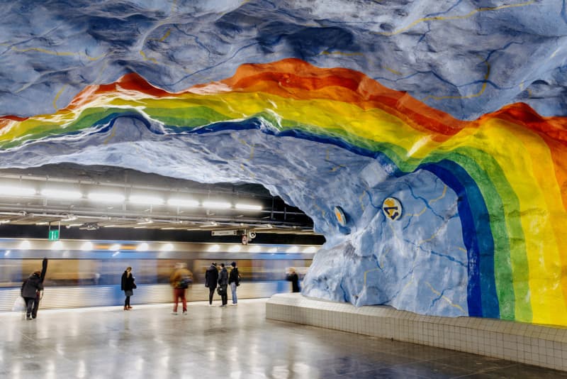 Rainbow in the Stockholm metro Vibrant rainbow mural painted across the blue rock wall of a metro station, with passengers waiting on the platform.