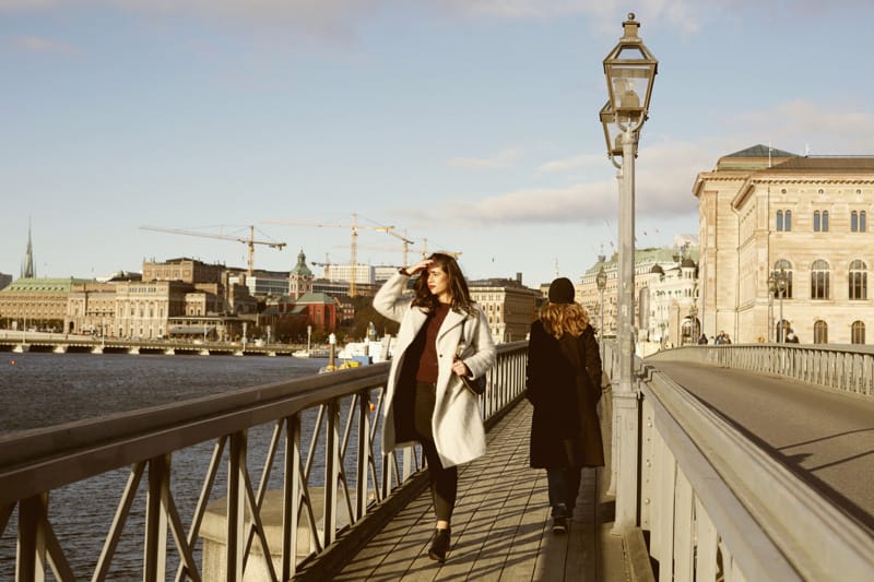 Two people walking on a bridge in an urban waterfront setting with historic buildings and construction cranes in the background during daytime.