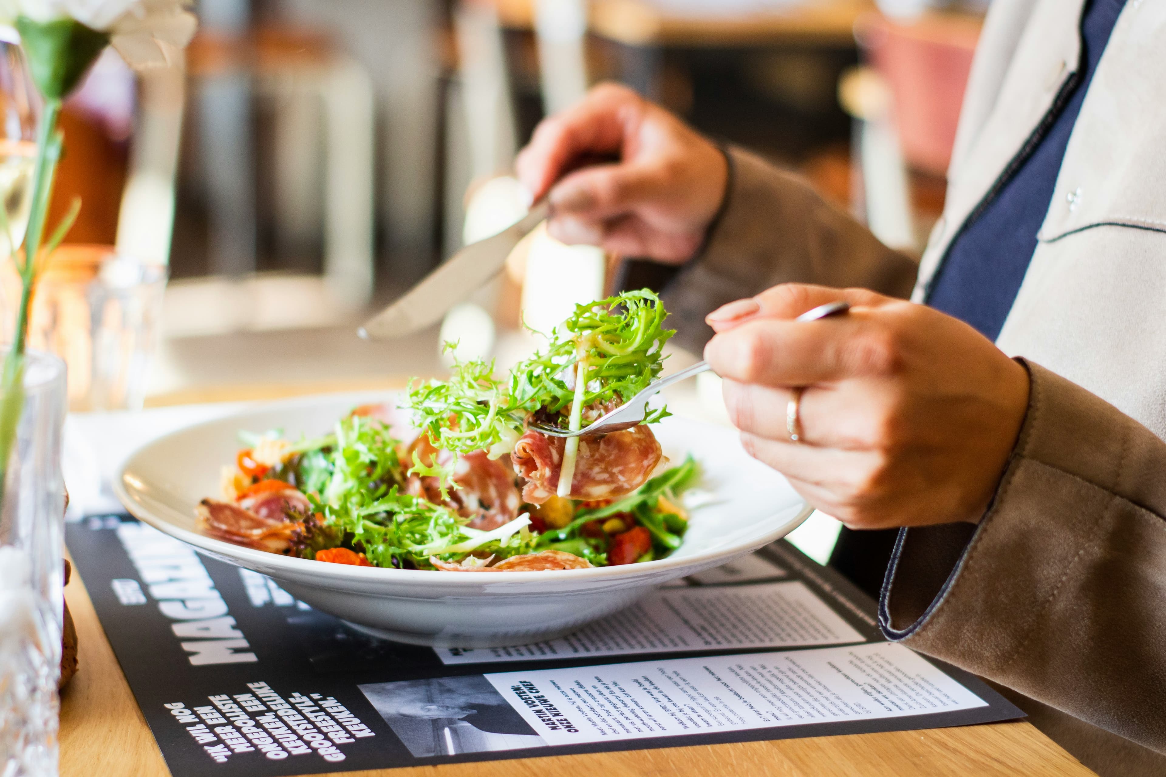 En person äter en färsk sallad med grönsaker och kött ur en vit skål på en restaurang i Stockholm, med varma trätoner i bakgrunden.