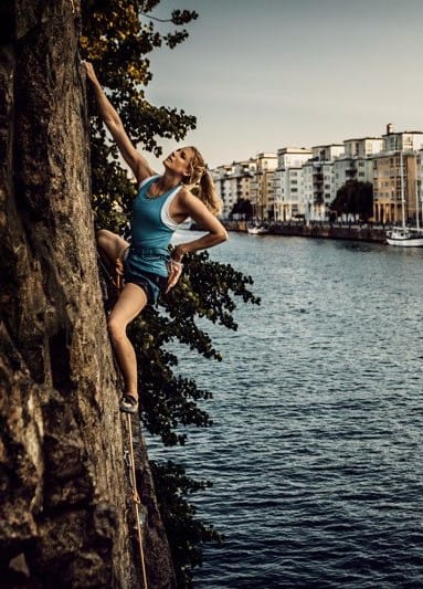 A person climbs a cliff face beside the water in an urban setting, with boats and modern buildings in the background.