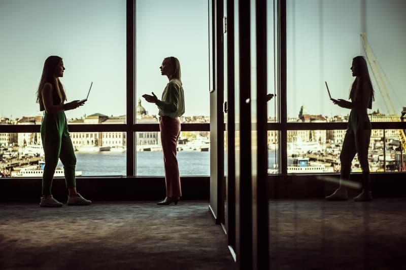 Two people stand in a glass-walled office corridor overlooking a city harbor, talking while one holds a laptop, with their silhouettes and reflections visible in the windows.