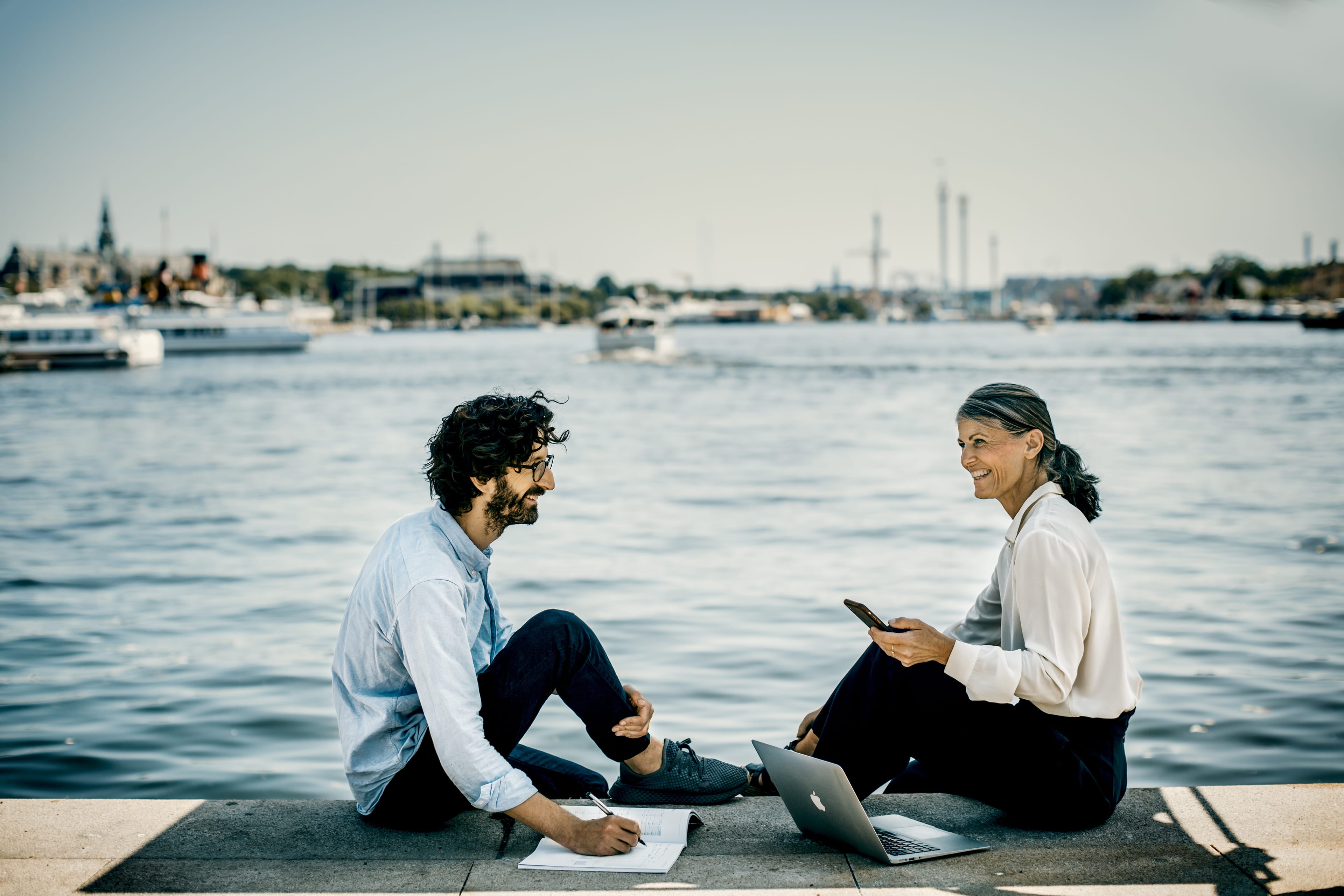 Two people sitting on a quay by the water with a notebook, phone, and laptop, talking in the sunlight.