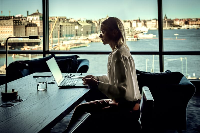 A person sitting at a table working on a laptop indoors, with large windows and a view of a harbor and cityscape in the background.