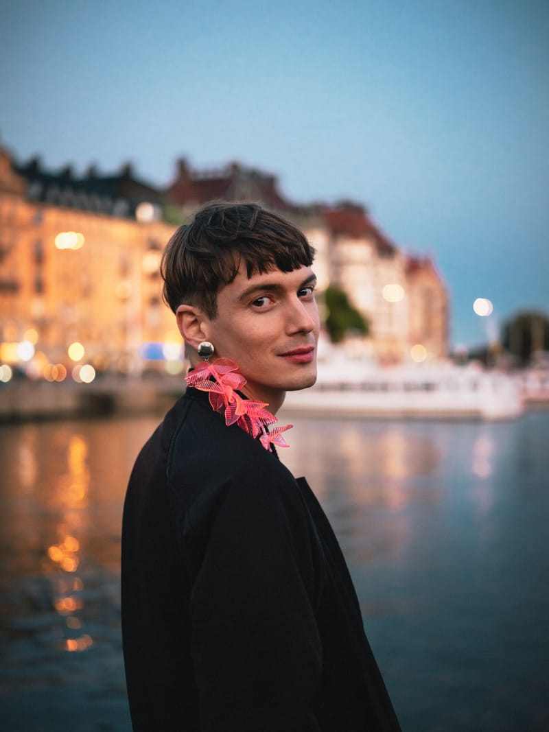 Person wearing a pink floral accessory around their neck standing by the waterfront at dusk with lit-up buildings in the background.