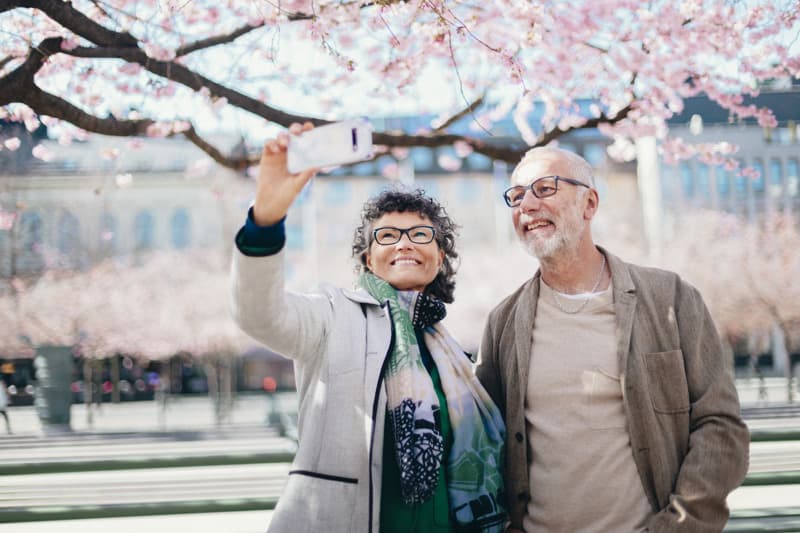 Två vuxna ler och tar en selfie med en mobiltelefon medan de står under blommande körsbärsträd i en stadspark.