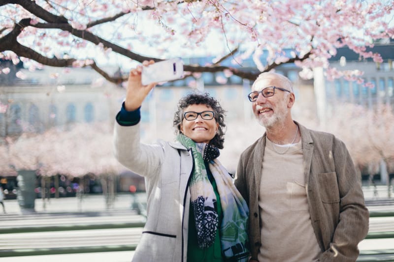 Two adults smiling and taking a selfie with a smartphone while standing under blooming cherry trees in an urban park.