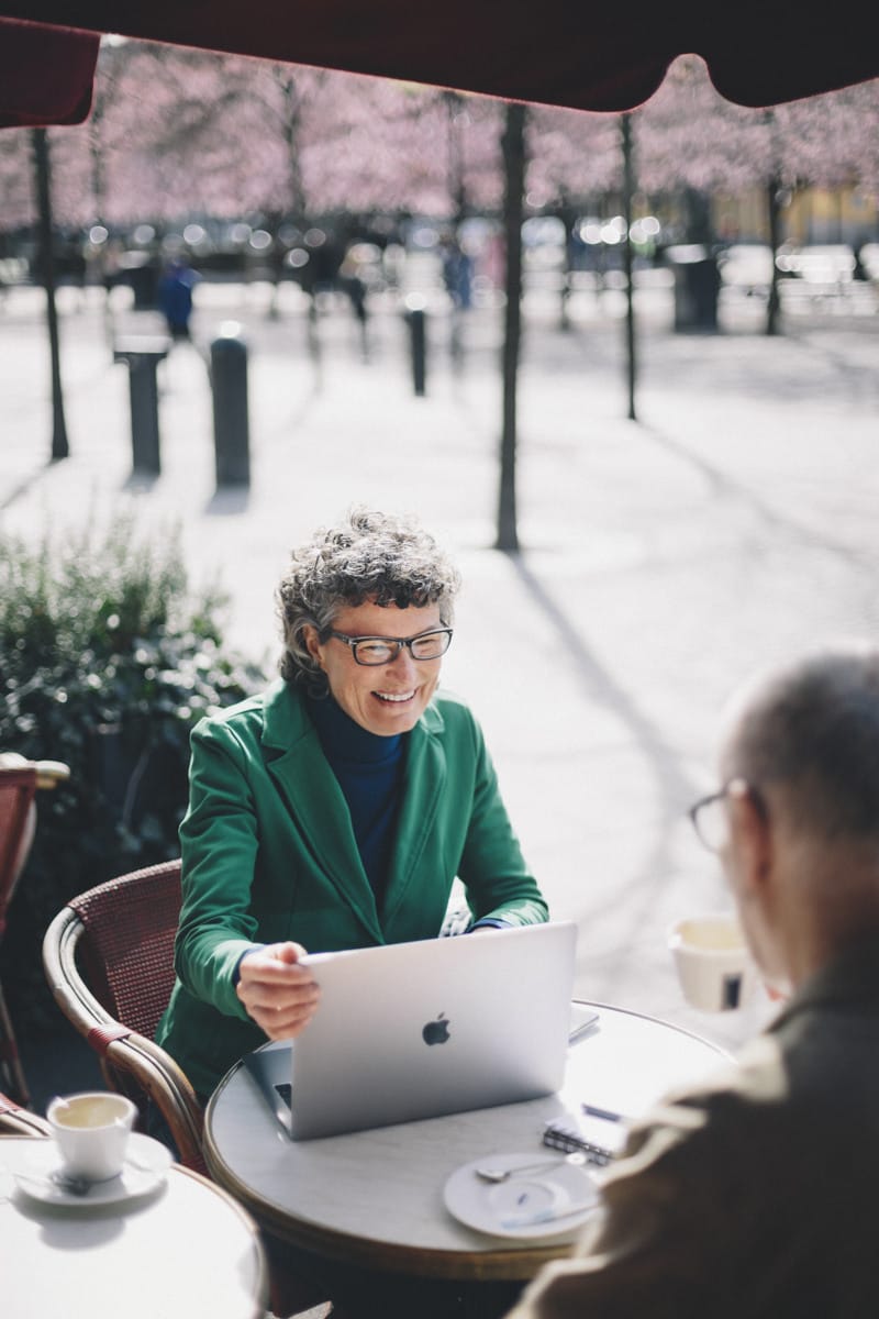 Person in a green blazer sitting at an outdoor café with a laptop, smiling while talking to a colleague, with blooming trees in the background.
