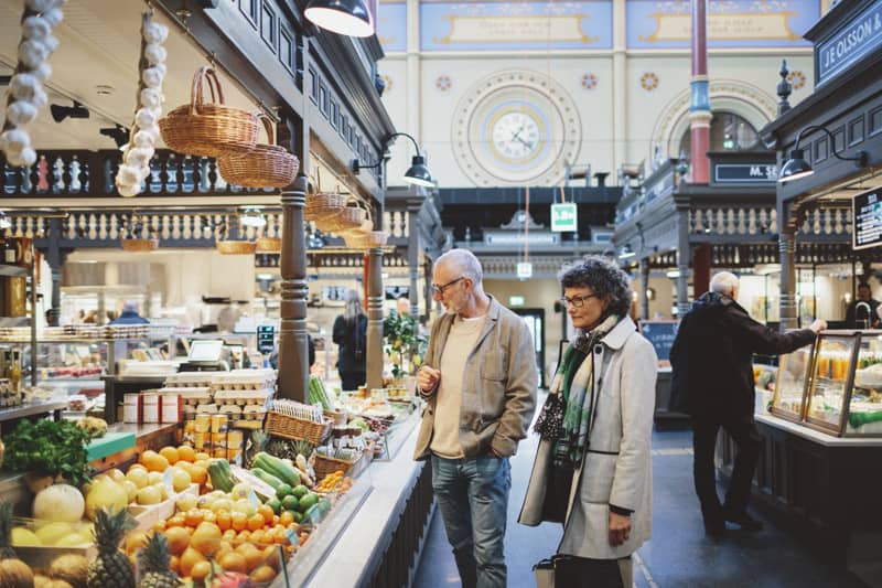 Browsing fresh produce in the market hall Two adults look at a colorful display of fruit and vegetables inside an ornate indoor market hall, with hanging baskets and a large decorative clock in the background.