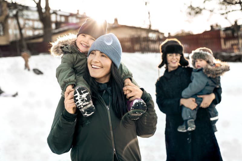 Winter play in the sunshine An adult smiles while carrying a laughing child on their shoulders in a snowy park, with another adult in the background holding a bundled-up child, all dressed in warm winter clothes.