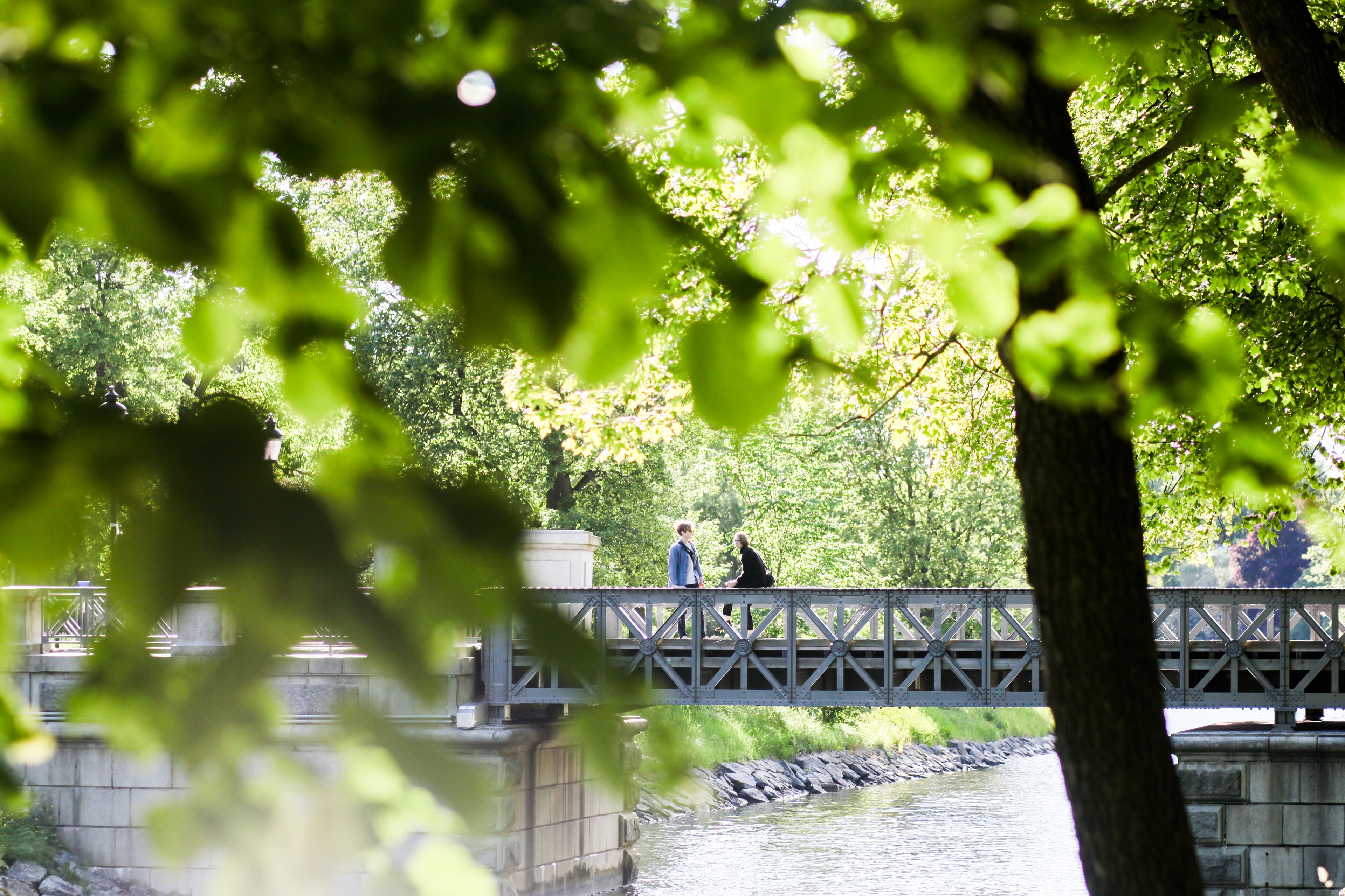 Two people stand talking on a footbridge over a canal, framed by lush green trees in a park.