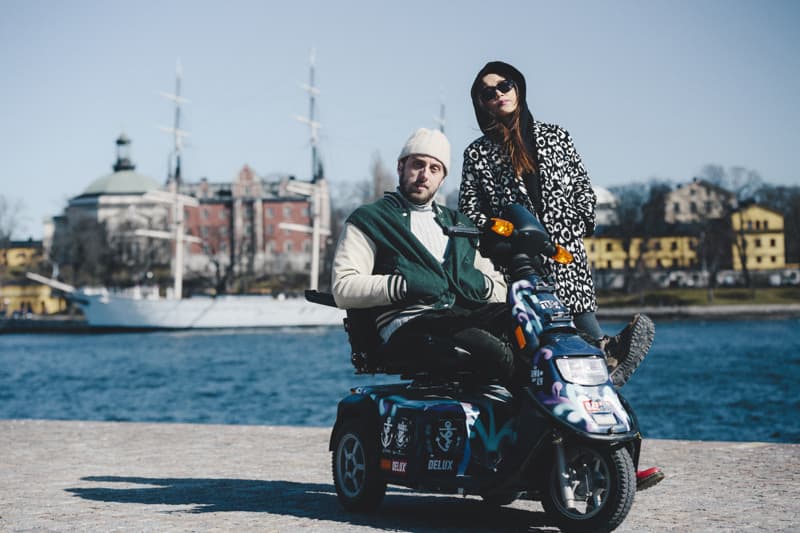 A person sits on a decorated mobility scooter beside a friend standing next to them, both wearing winter clothes, with water, a white sailing ship and colorful buildings in the background.