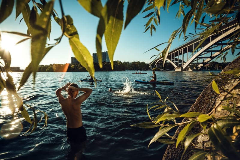 People swimming and paddleboarding in the evening sun near a bridge, seen through overhanging green leaves by the water.