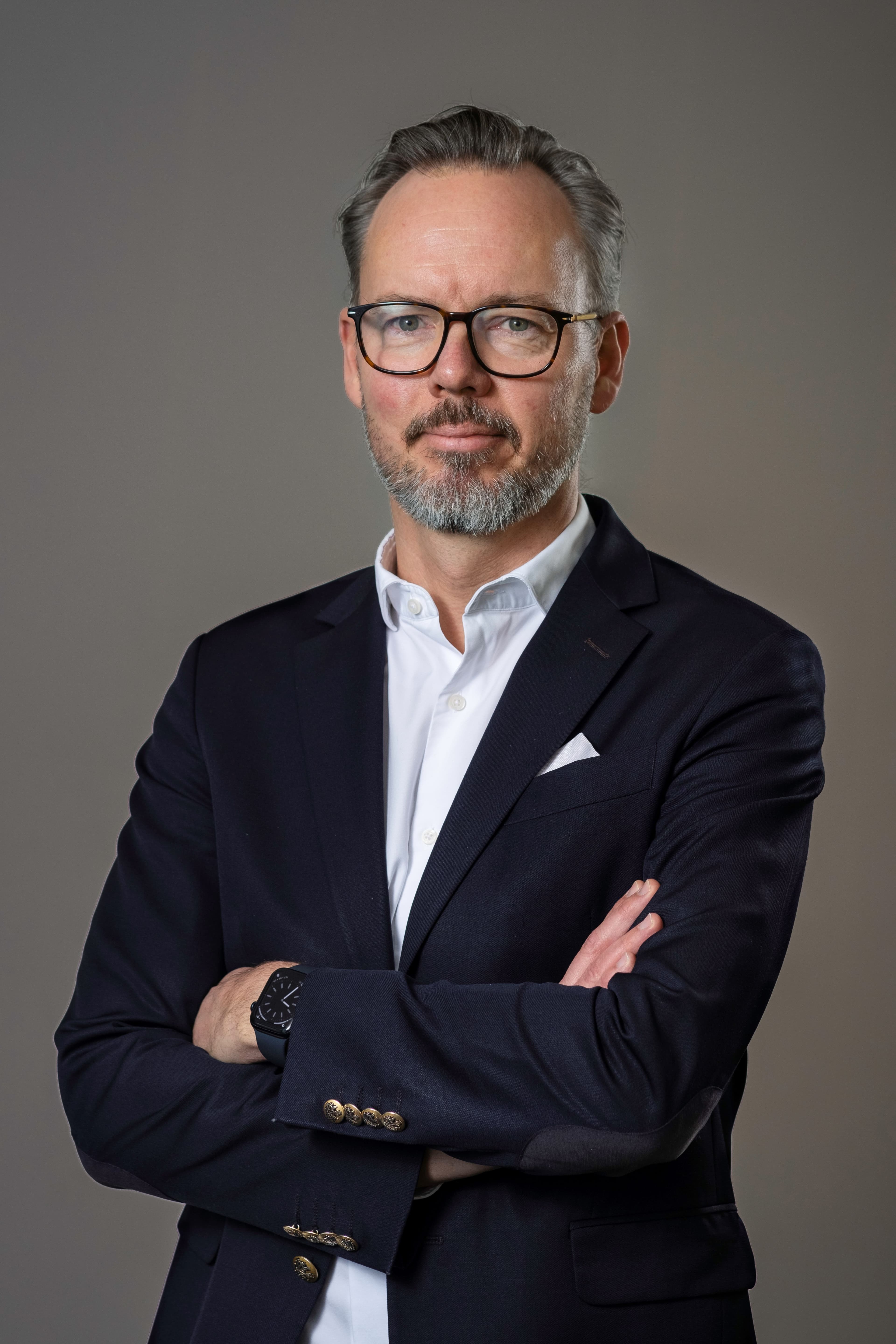 Middle-aged man with glasses and a beard wearing a dark suit and white shirt, standing with arms crossed against a neutral background.