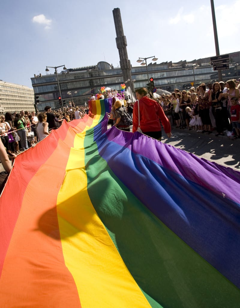 A large rainbow flag is carried by participants during a pride parade in an urban setting, surrounded by spectators and modern buildings.