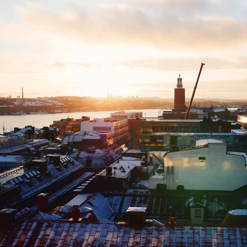 A warm winter sunset over central Stockholm, with snow-dusted rooftops in the foreground, the water and skyline in the distance, and the City Hall tower silhouetted against the glowing sky.