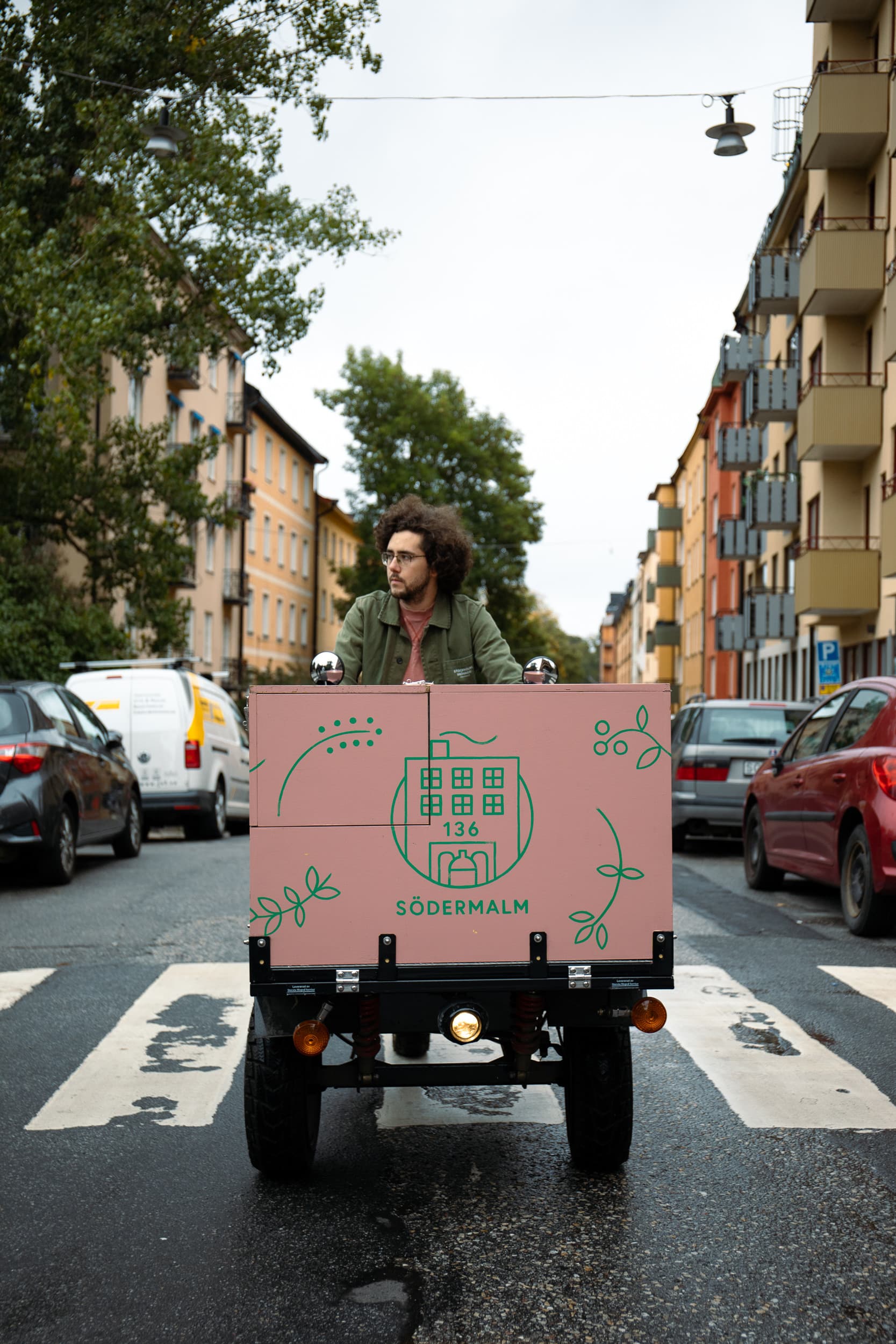 Person kör ett litet lastfordon med rosa låda märkt ”Södermalm” mitt på en stadsgata med bostadshus och parkerade bilar.