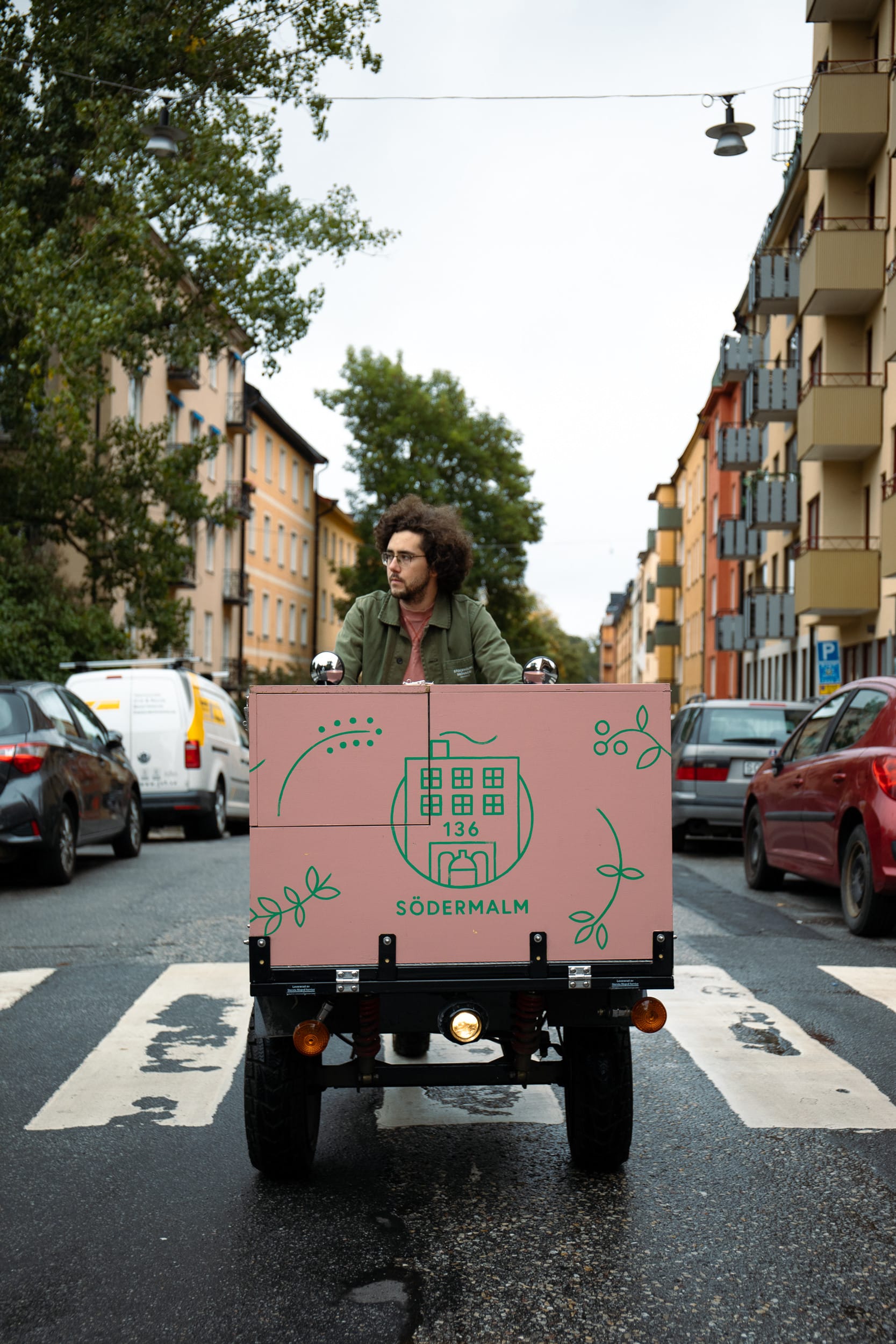 Person driving a small cargo vehicle with a pink box labeled “Södermalm” in the middle of a city street lined with apartment buildings and parked cars.