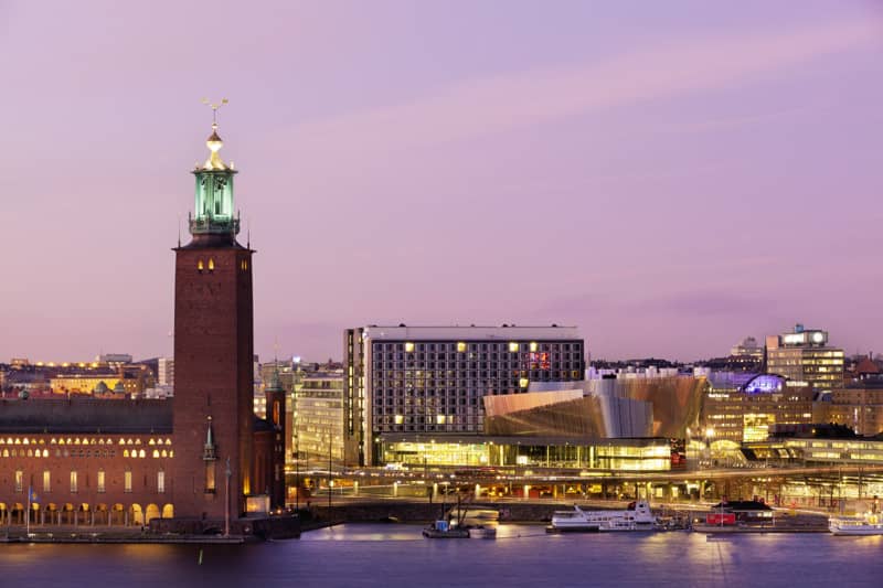 City Hall and Waterfront in evening light Stockholm City Hall’s brick tower and the modern waterfront congress building are lit up along the quayside at dusk, with glowing windows, boats in the water and a purple sky above.