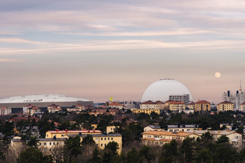 Southern Stockholm with Avicii Arena at dusk View over a residential area in southern Stockholm with a large white spherical arena and a flat-roofed stadium on the horizon, under a pastel evening sky with the moon rising.